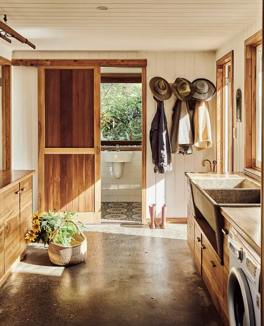 Rustic mudroom laundry room combo with warm wood cabinets, deep utility sink, wall hooks, hats, coats, and a front-loading washer.