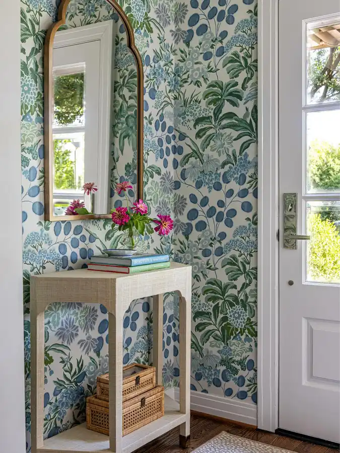 Entryway table styled with botanical wallpaper, a tall scalloped mirror, colorful flowers, stacked books, and woven storage baskets.