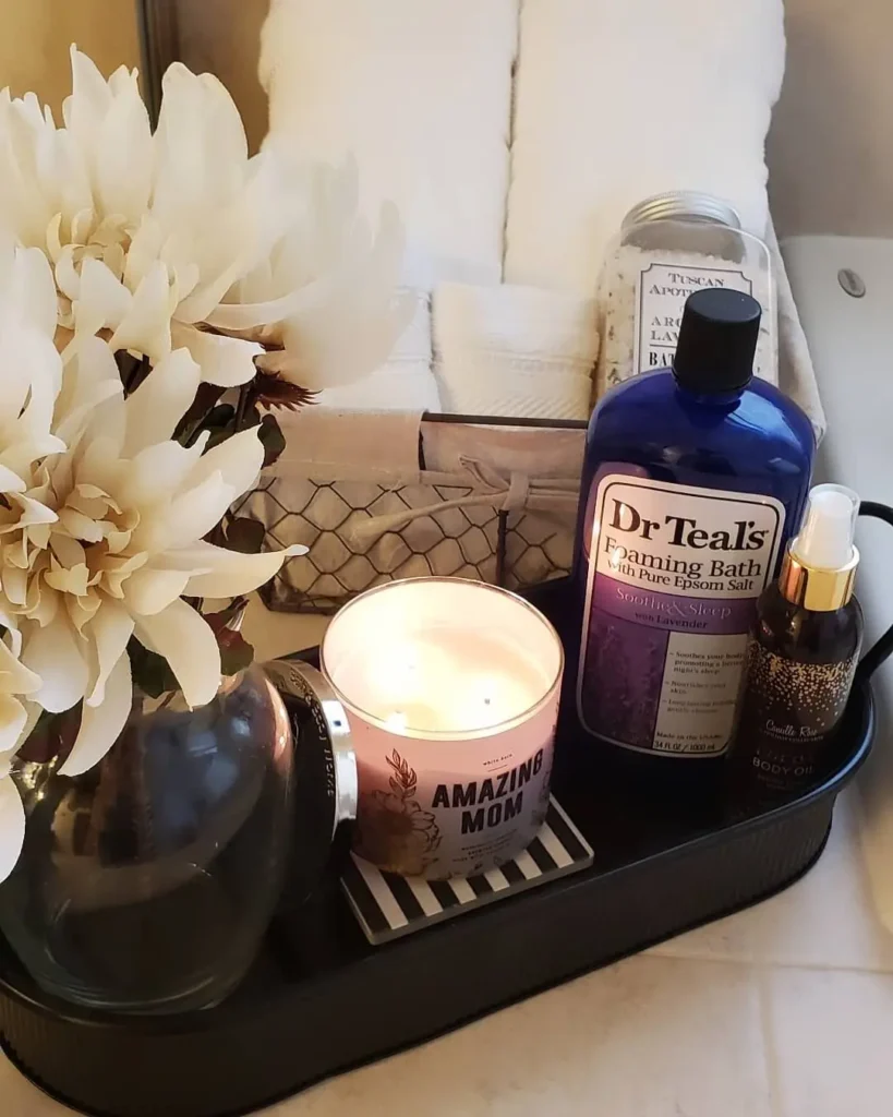 Black bathroom tray holding a candle, bath products, body oil and rolled white towels beside ivory flowers.