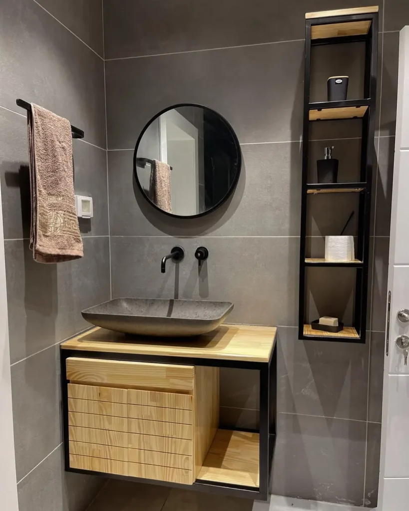 Modern bathroom vanity with a wide vessel sink, warm wood counter, black faucet, round mirror, and slim wall shelves.