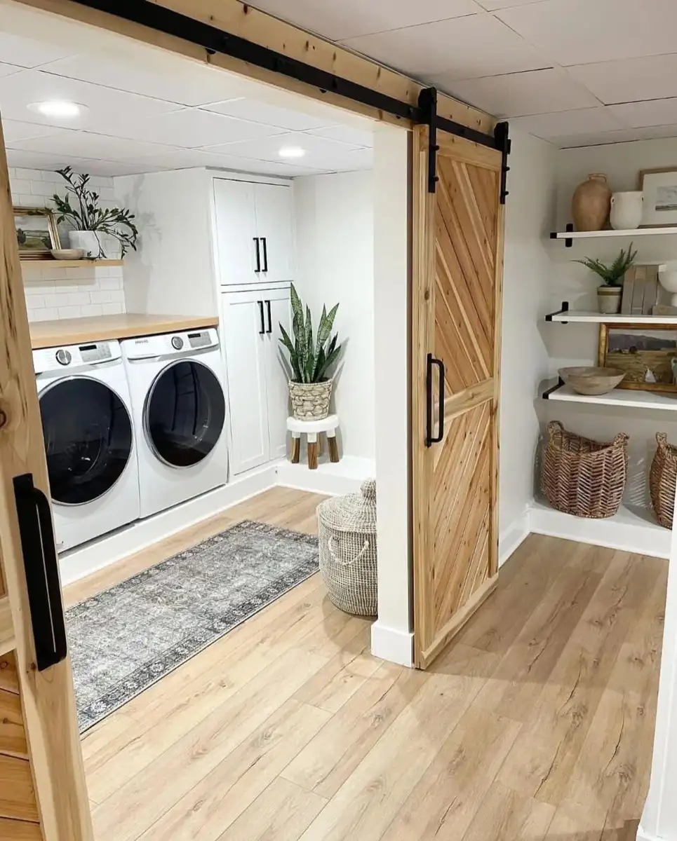 Bright mudroom laundry room with white cabinets, side-by-side washer and dryer, folding counter, open shelves, and sliding wood barn doors.