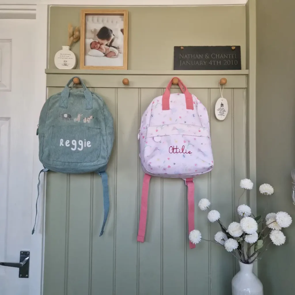 Two children’s backpacks hanging from wall hooks beneath a narrow display shelf in a sage green entryway.