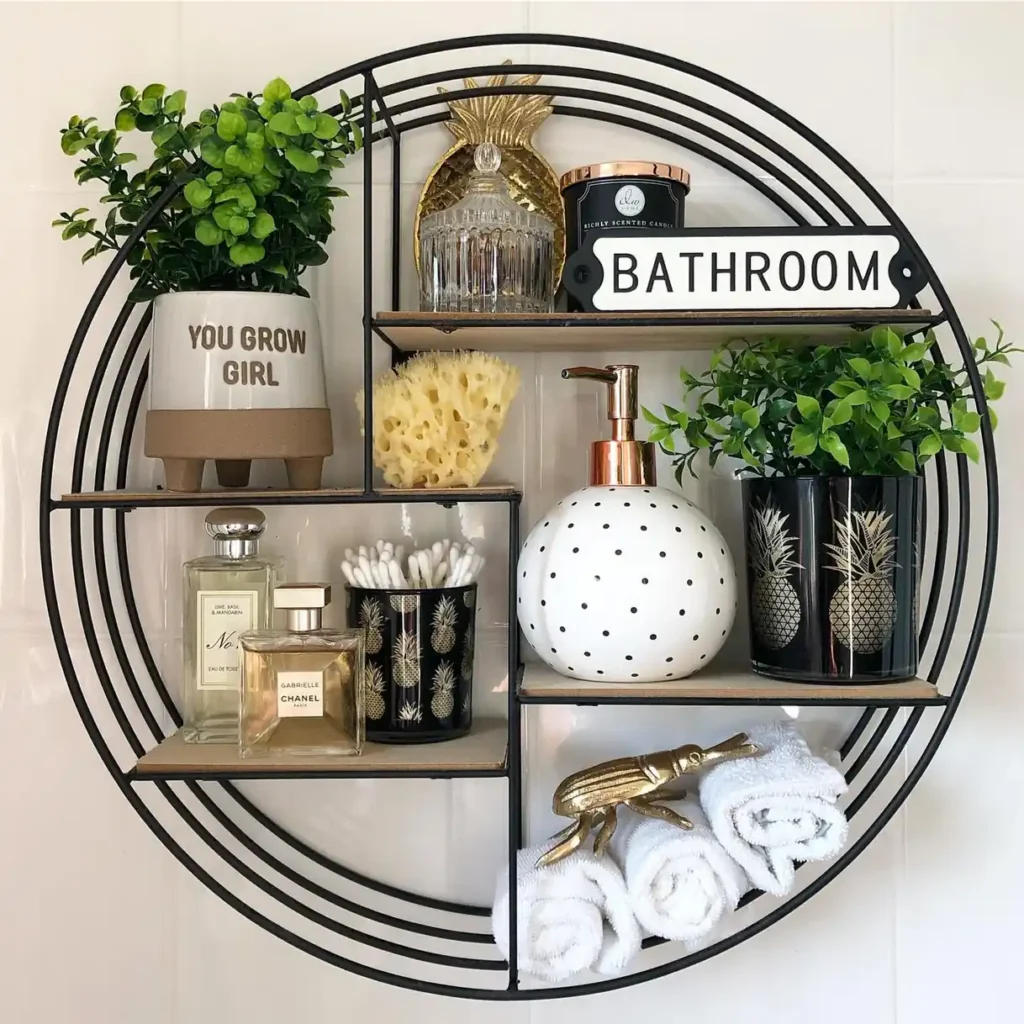 Round black bathroom wall shelf styled with greenery, rolled towels, jars, soap dispenser, and gold accents.