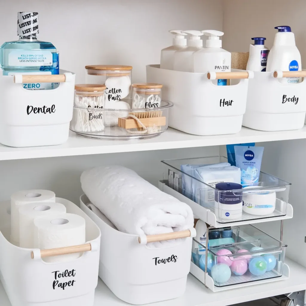 Bathroom closet organization with labeled bins, clear drawers, glass jars, folded towels, and toilet paper storage.