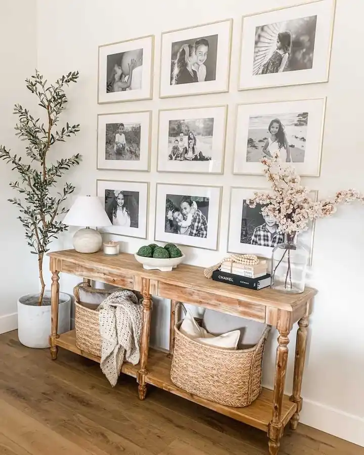 Warm wood entryway console beneath a grid of black and white family photos, styled with a lamp, flowers, books, and woven baskets.