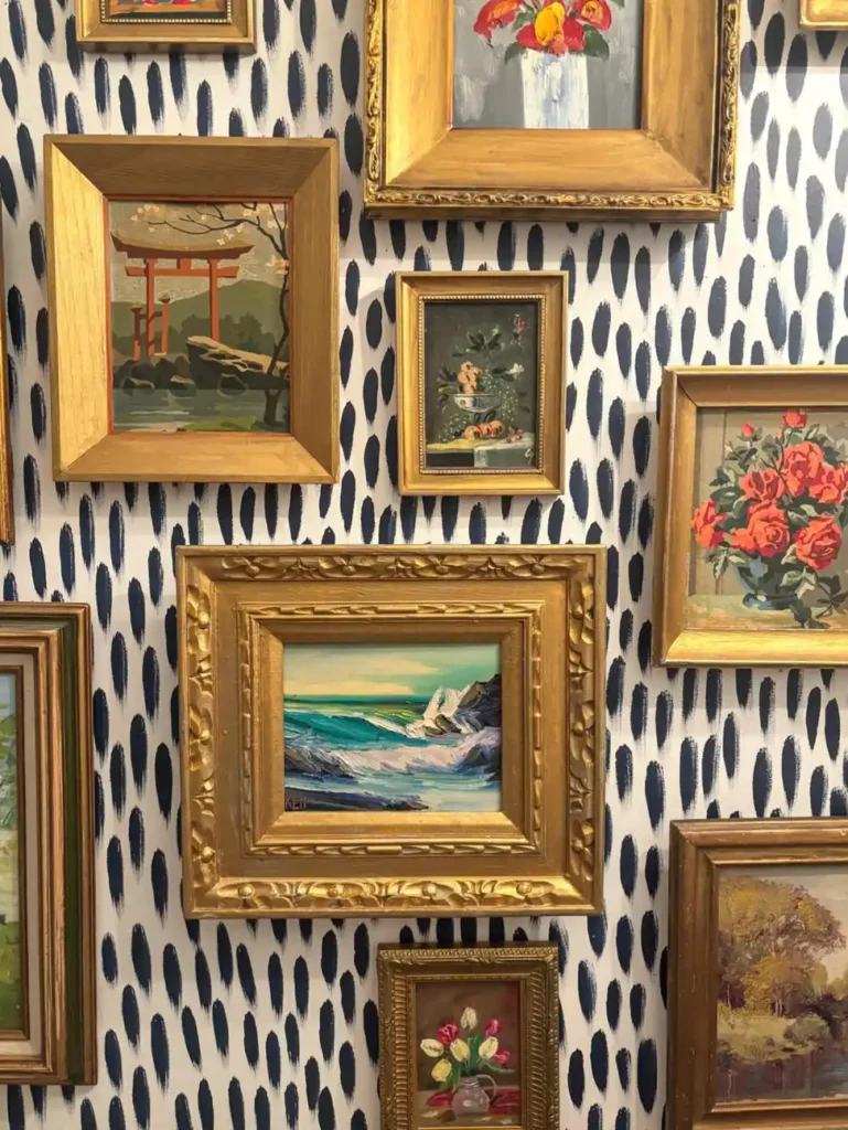 Eclectic gallery wall with gold framed floral paintings, landscapes, and seascapes over patterned wallpaper.
