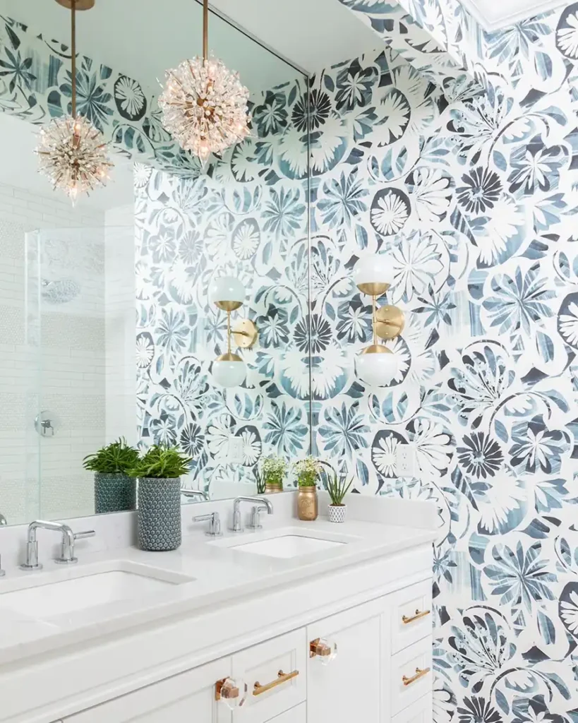 Bright bathroom featuring blue floral wallpaper, a white vanity, tall mirror, brass lighting, and small green plants.