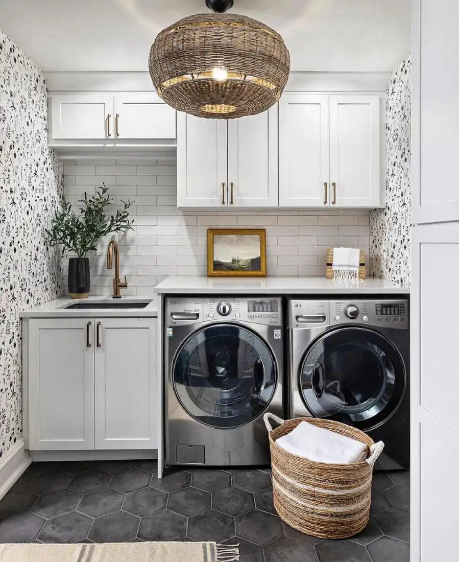 Stylish laundry room with floral wallpaper, white cabinets, utility sink, dark hex tile floor, wicker basket, and front-loading washer and dryer.