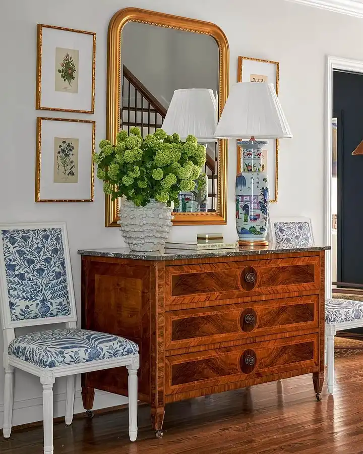 Traditional wood dresser used as an entryway console with a large gold mirror, patterned lamp, green floral arrangement, and botanical wall art.