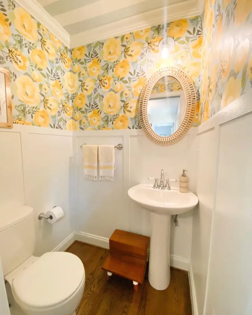 Small bathroom with yellow floral wallpaper, pale paneled walls, a pedestal sink, and an oval woven mirror.