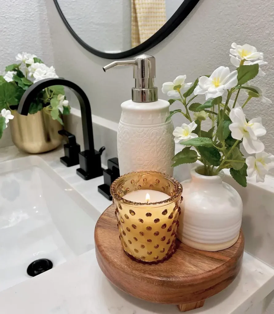 Raised round wooden tray holding an amber candle and small white flower vase beside a soap dispenser.