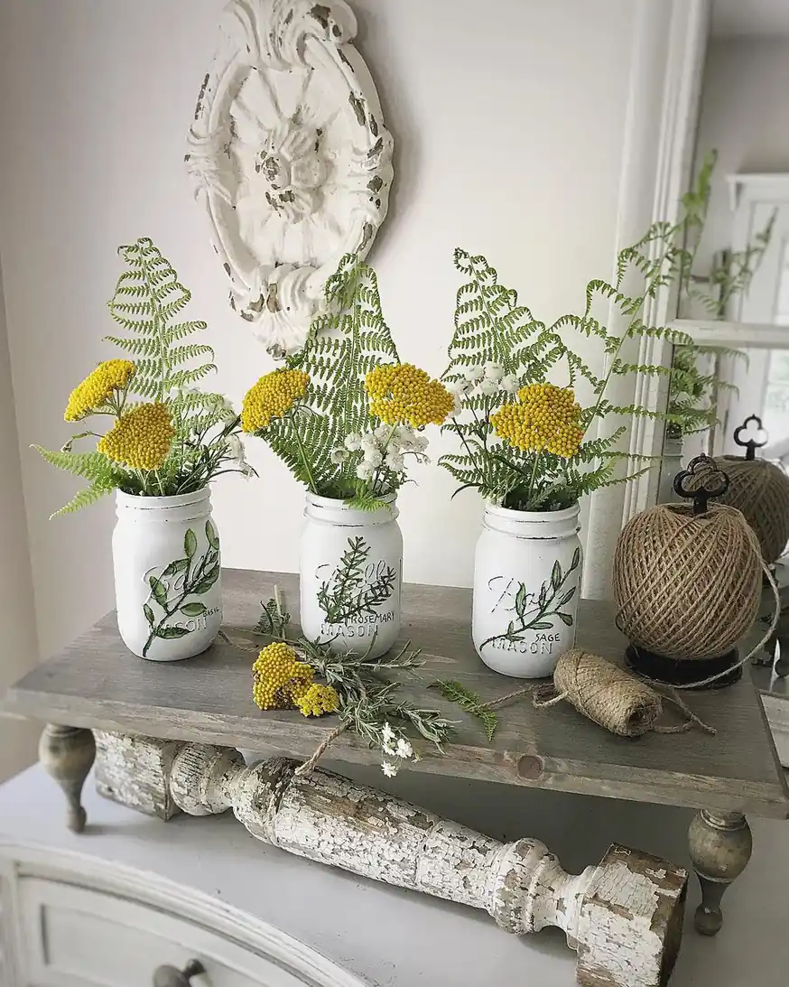 Three white painted mason jars with yellow flowers and greenery displayed on a wooden stand