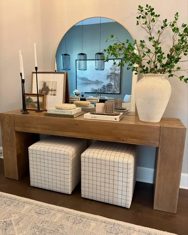 Rustic wood entryway console with two upholstered stools underneath, a round mirror, leafy branches, candlesticks, books, and leaning artwork.