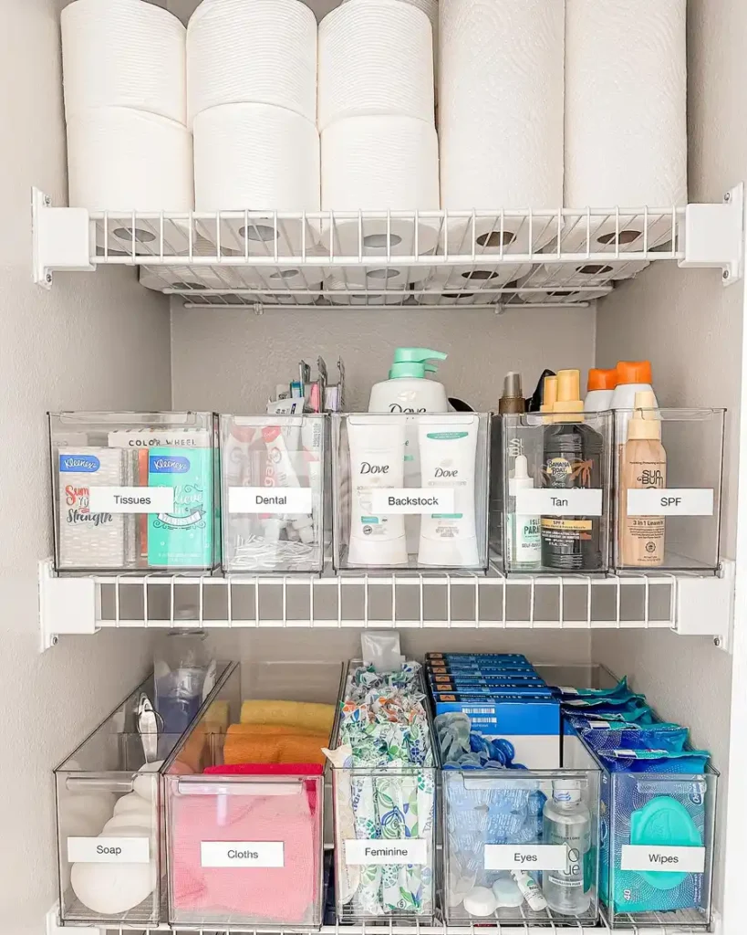 Bathroom closet with clear labeled bins arranged on wire shelves for toiletries, soap, towels, wipes, and extra supplies.