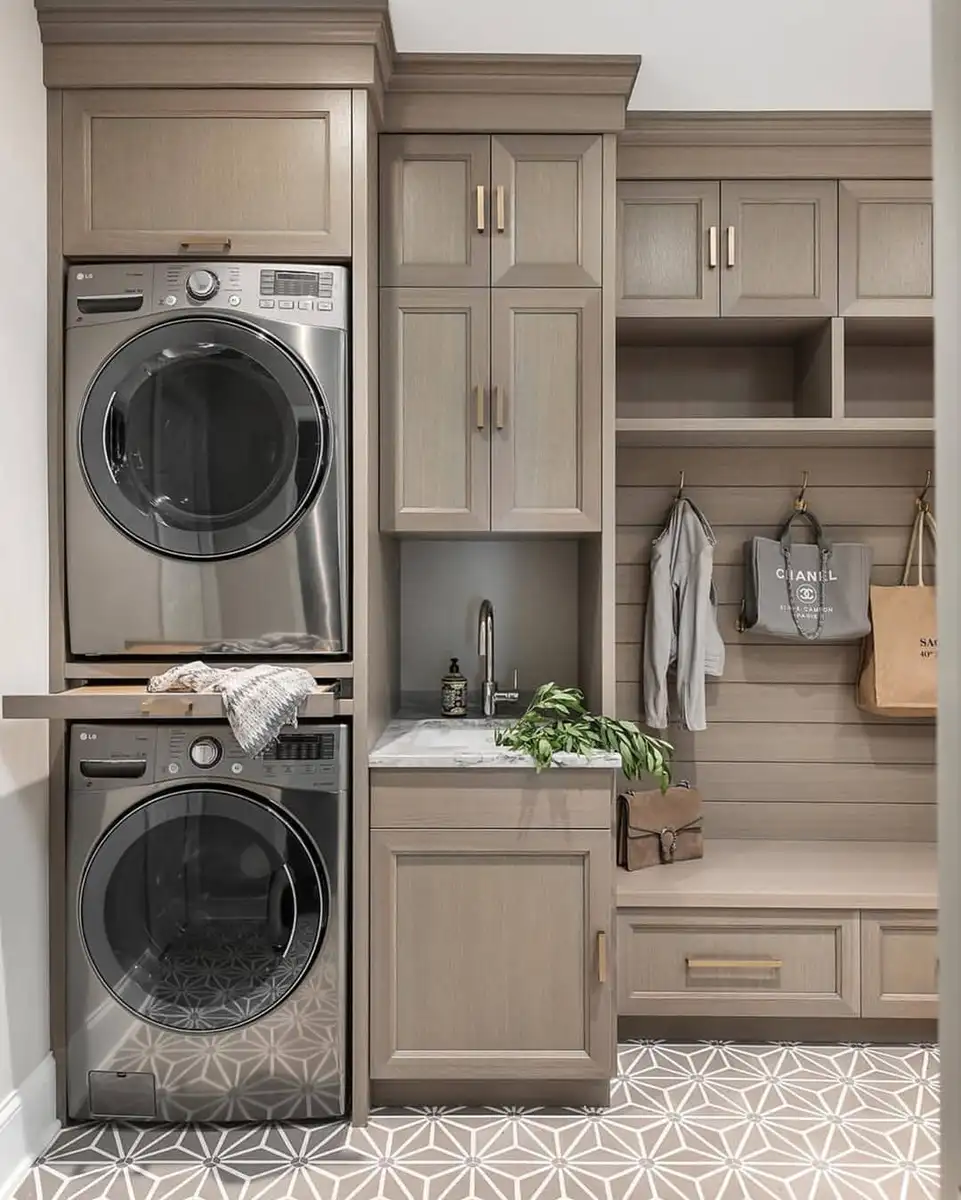 Small mudroom laundry room combo with stacked washer and dryer, utility sink, tall cabinets, storage bench, wall hooks, and patterned tile floor.
