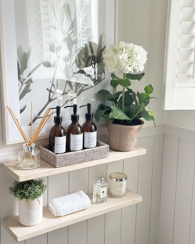 Pale wood bathroom shelves styled with pump bottles in a woven tray, white towels, greenery, candle, and diffuser.