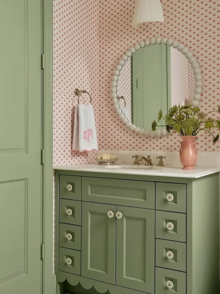 Sage green bathroom vanity with a white sink, soft pink accents, floral wallpaper, leafy arrangement, and round textured mirror.