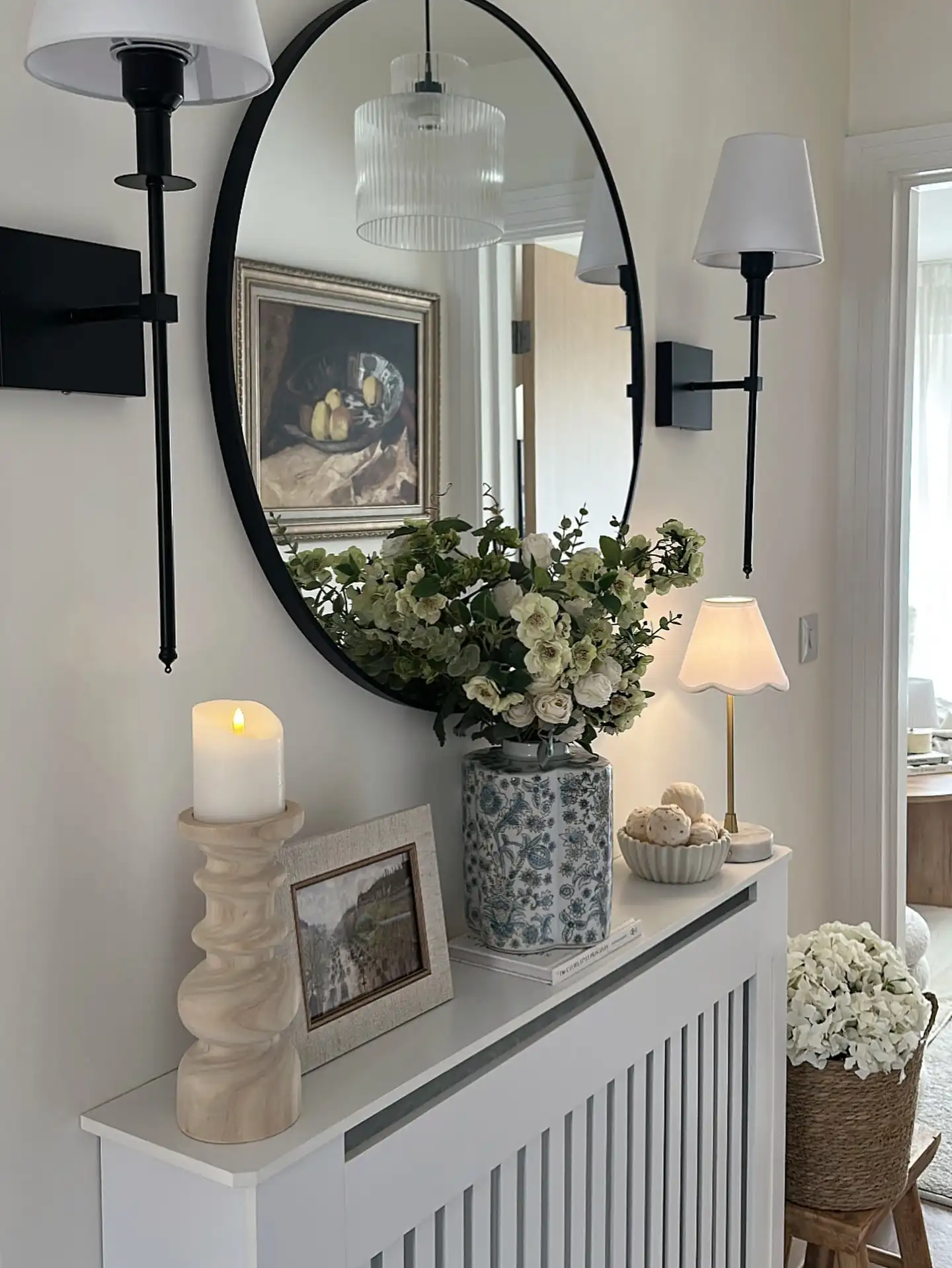 Elegant hallway console with large black framed mirror, white flowers, table lamp, sconces, and neutral decor
