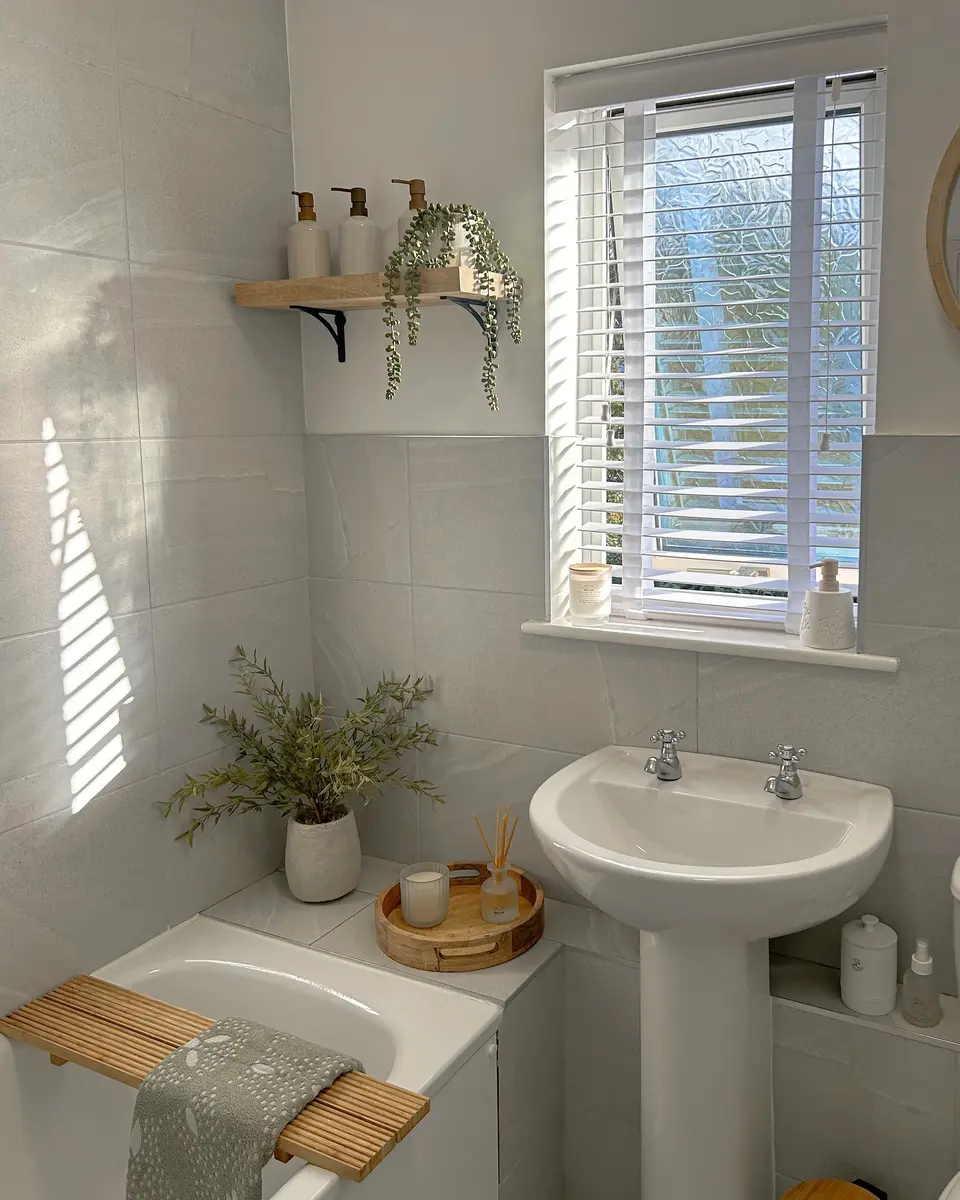 Bright compact bathroom with a pedestal sink, gray wall tiles, window blinds, plants, and a wooden bath caddy.