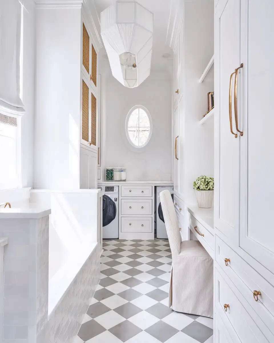 Bright white mudroom laundry room galley with tall built-in cabinets, bench seating, side-by-side washer and dryer, brass hardware, and checkered floor.