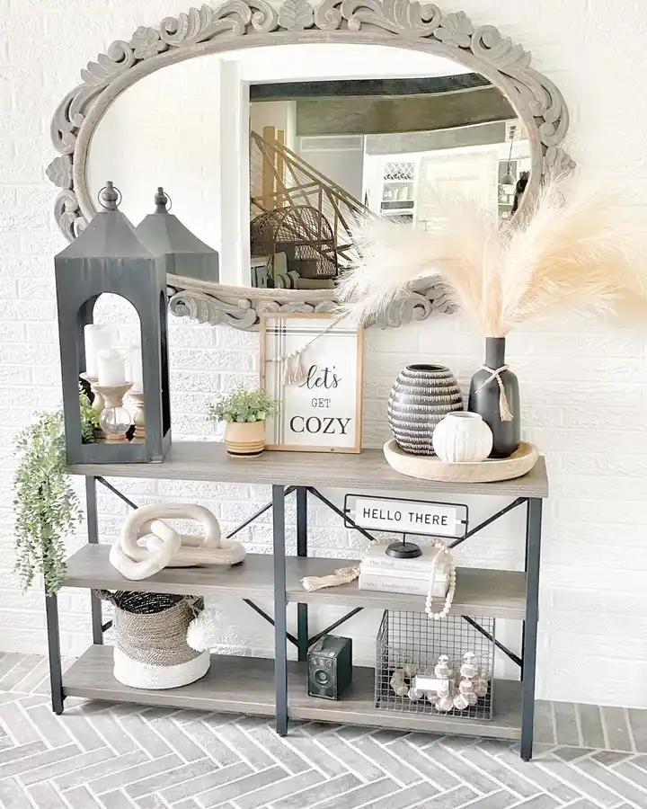 Farmhouse entryway console with a carved mirror, black metal frame, pampas grass, lantern, signs, pottery, books, and neutral accessories.