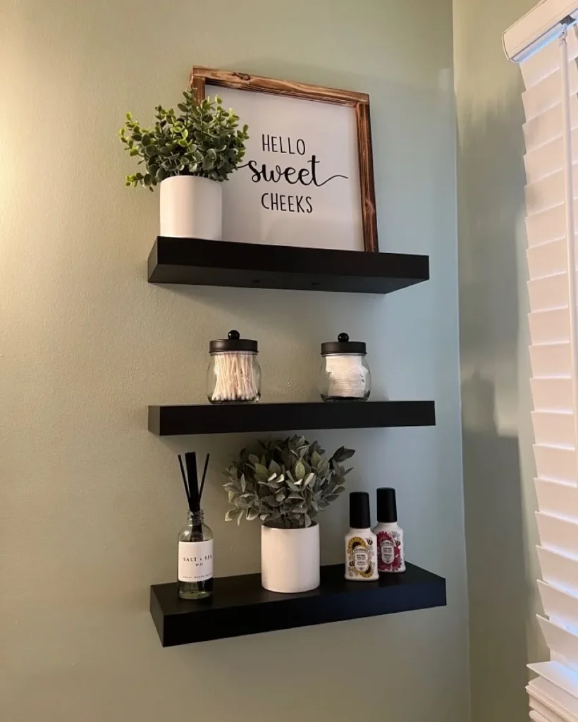 Black floating bathroom shelves with framed sign, cotton jars, greenery, reed diffuser, and small toiletry bottles.
