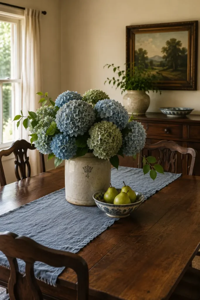 Blue and green hydrangeas in a stoneware crock beside a patterned bowl of pears on a blue runner.