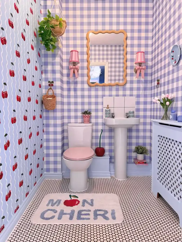 Lavender gingham bathroom with cherry-print wallpaper, a pink toilet seat, bow wall lights, and a wavy wooden mirror.
