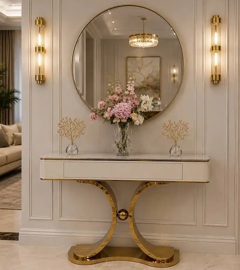 Elegant cream entryway console with gold trim, large round mirror, matching wall sconces, floral centerpiece, and small glass accents.