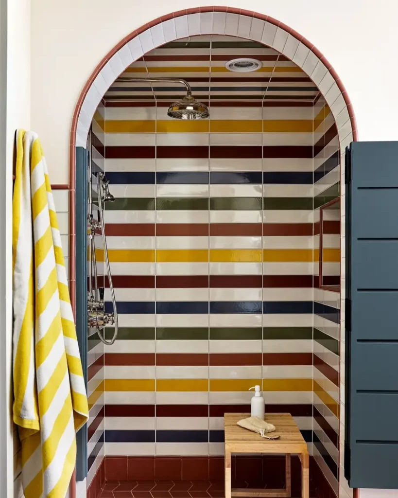 Arched shower covered in colorful horizontal tile stripes with dark blue doors, a wooden stool, and a yellow towel.
