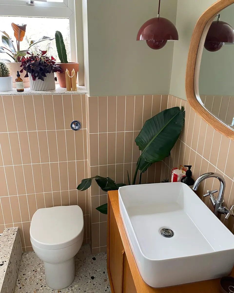 Small peach and sage bathroom with vertical wall tiles, a vessel sink, round wood mirror, and leafy plants.