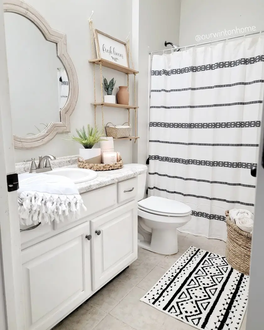 Small farmhouse bathroom with rope-hung wood shelves, patterned shower curtain, woven laundry basket, white vanity, and decorative mirror