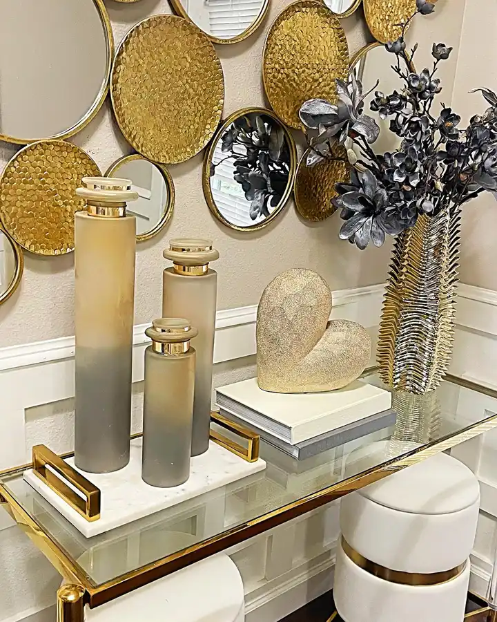 Glass entryway console styled with gold wall mirrors, metallic decor, tall decorative bottles, stacked books, dark branches, and stools underneath.