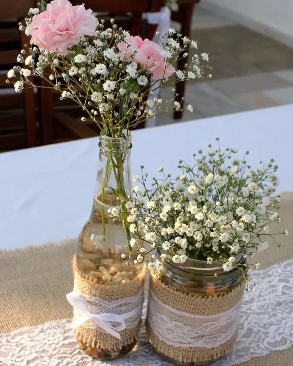 Lace and burlap wrapped mason jar with baby’s breath beside a glass bottle filled with pink flowers