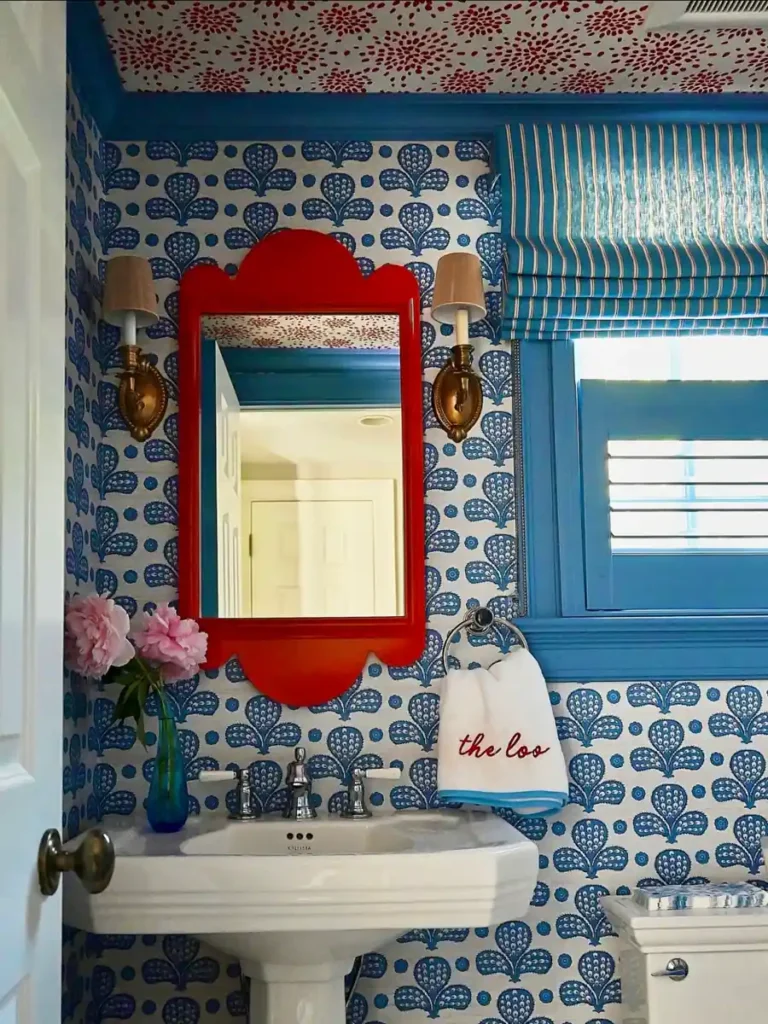 Blue floral powder room with a red scalloped mirror, brass sconces, striped window shade, and white pedestal sink.