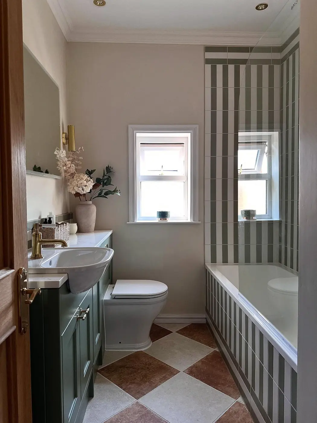 Narrow bathroom with a green vanity, striped shower tiles, a glass screen, diamond flooring, and a bright far window.
