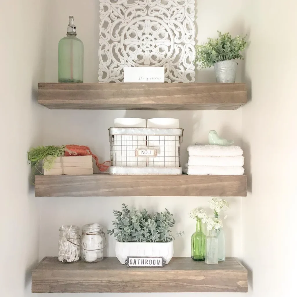 Rustic floating bathroom shelves with folded white towels, wire basket, glass jars, greenery, and decorative accents.