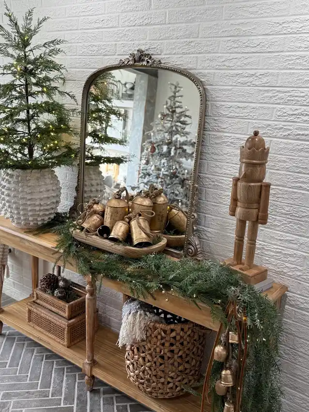 Rustic Christmas entryway console with a small lit tree, evergreen garland, wooden nutcracker, antique-style bells, ornate mirror, and woven baskets.