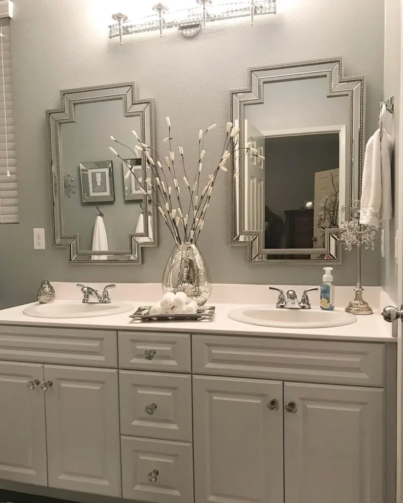 Glam double bathroom vanity with a tall silver vase of white branches and rolled towels arranged between two sinks.