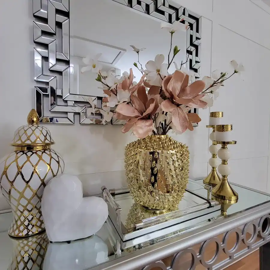 Glam mirrored entryway console with geometric mirror, blush flowers, gold candleholders, ginger jar, white sculpture, and metallic accents.