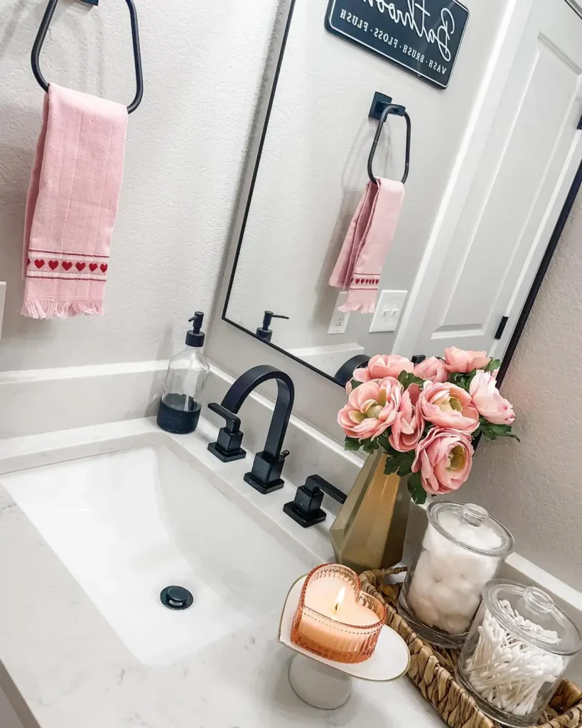 Bathroom counter with clear jars for cotton essentials, pink flowers, a heart shaped candle, woven basket, and pink towels.