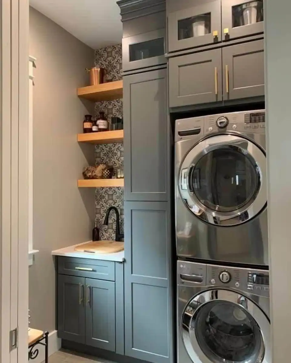 Compact laundry room with stacked washer and dryer, tall gray cabinet, utility sink, wood floating shelves, brass hardware, and patterned backsplash.