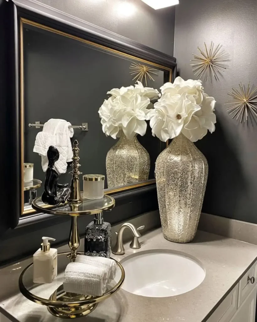 Dark glamorous bathroom vanity with oversized white flowers in a metallic vase, tiered towel stand, candle, and silver accents.