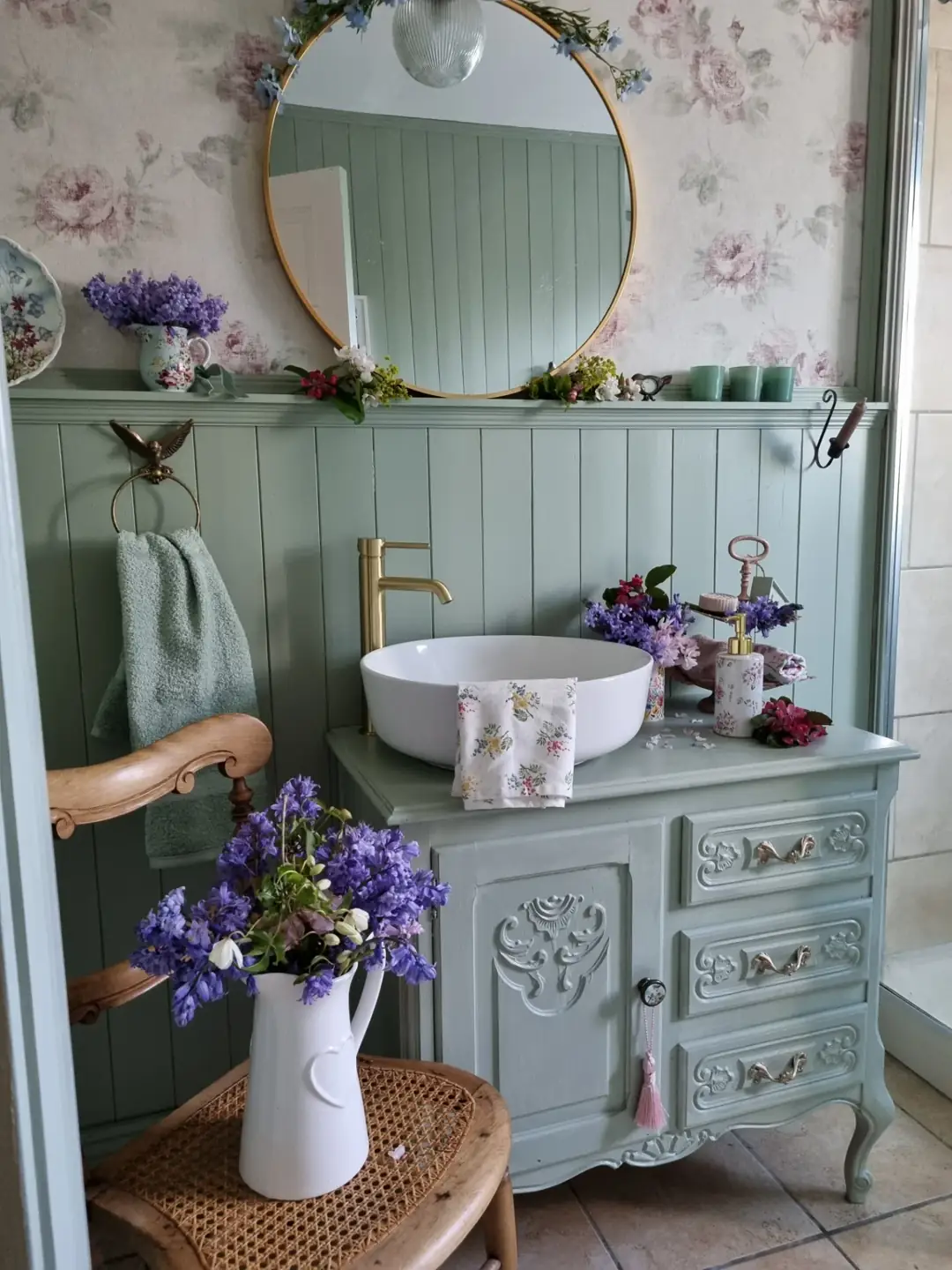 Elegant sage green bathroom with floral wall panels, white freestanding tub, brass faucet and vintage side tables