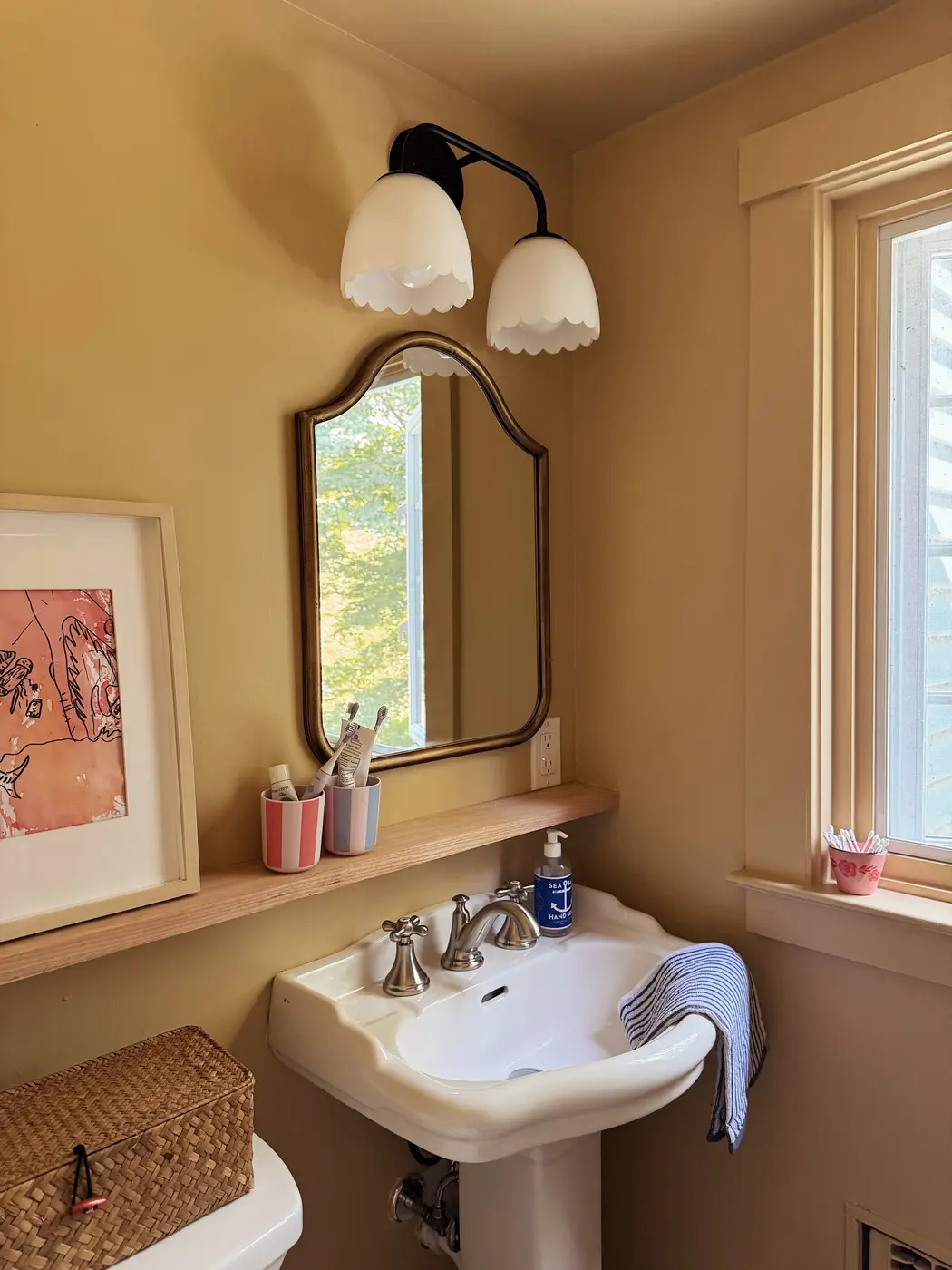 Warm yellow bathroom with a pedestal sink, arched mirror, narrow wooden ledge, wall light, and bright window.