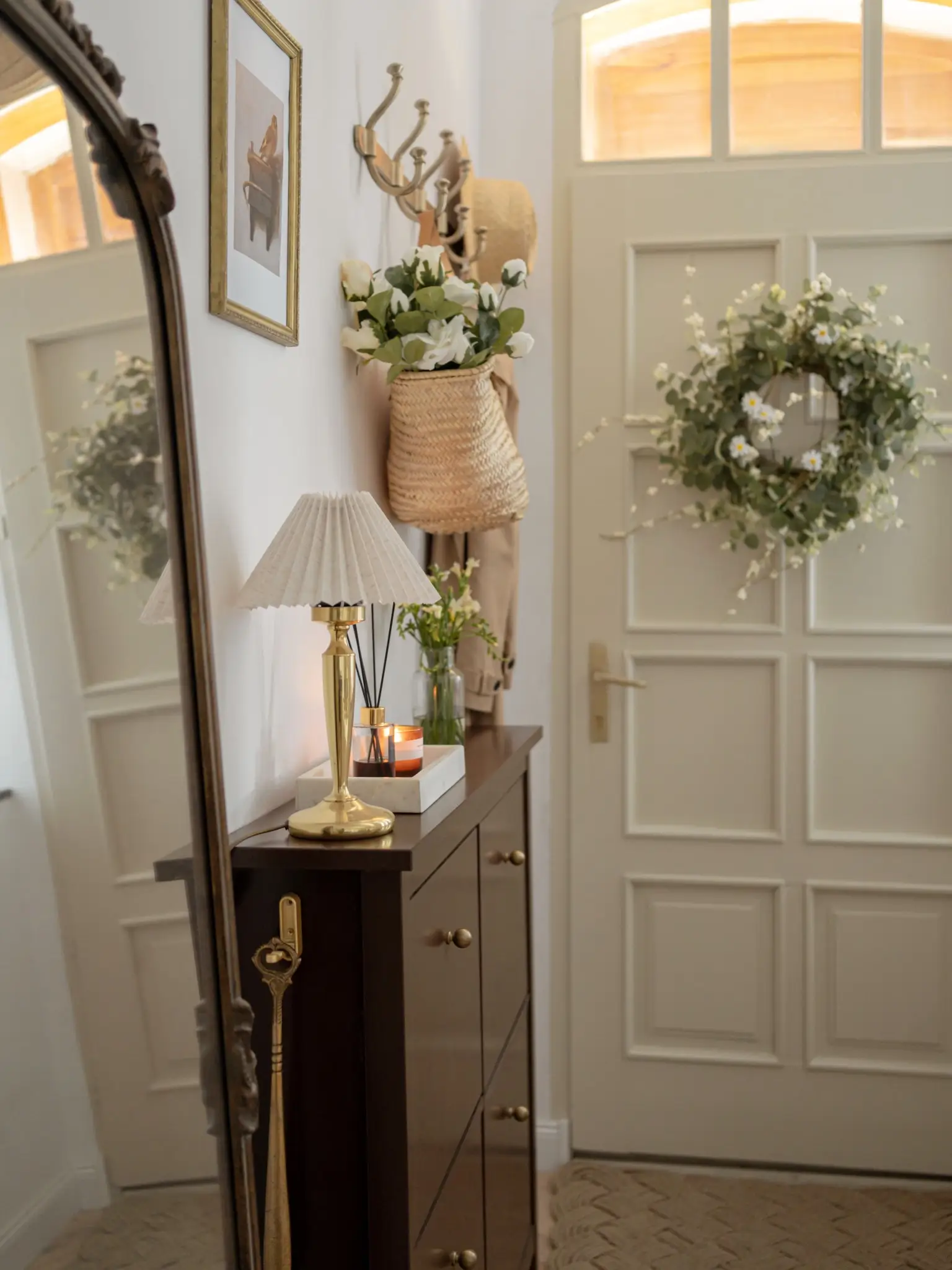Small entry hallway with tall leaning mirror, slim dark cabinet, brass lamp, wall hooks, wreath, and woven bag