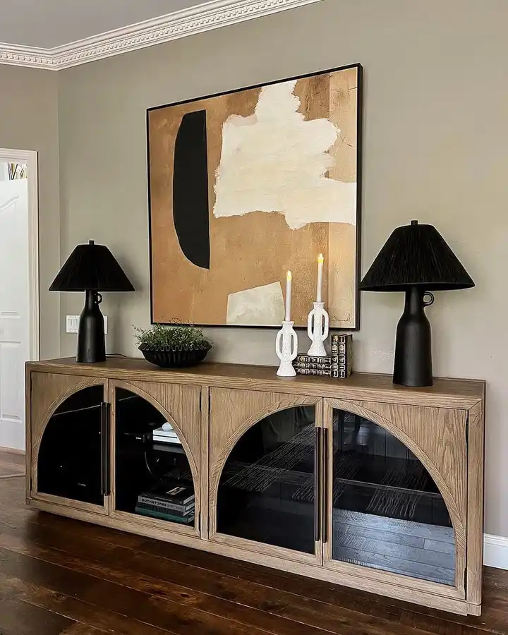 Warm wood entryway cabinet beneath oversized abstract art, styled with matching black lamps, white candleholders, greenery, and stacked books.