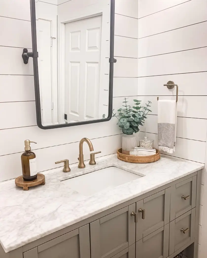 Modern farmhouse vanity with gray cabinetry, white shiplap, marble countertop, aged brass faucet, black mirror, and woven tray