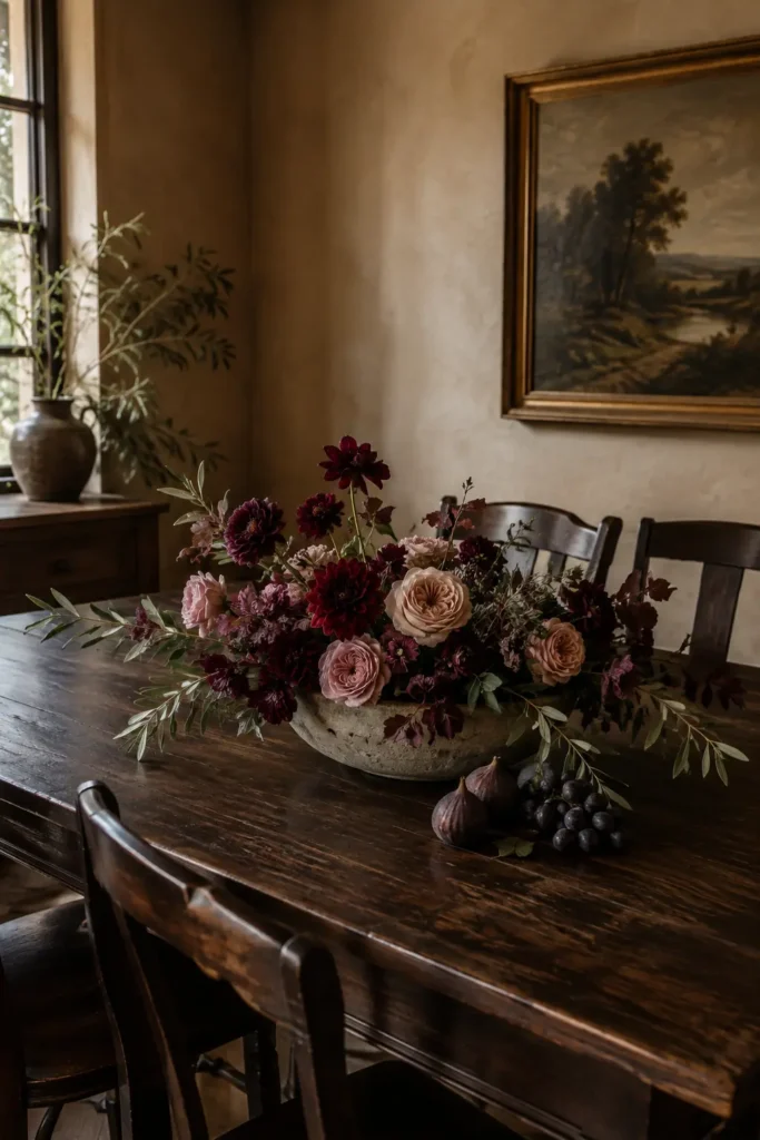 Burgundy flowers and blush roses in a low stone bowl with figs and dark grapes beside it.
