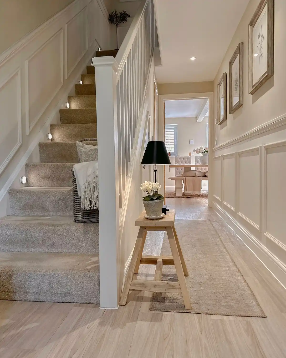 Light stair hallway with wall panel molding, narrow bench, black lamp, framed prints, and pale runner